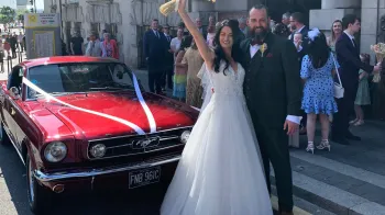 Bride and Groom cheering in front of a classic Ford Mustang in Cherry Red. Wedding guests can be seen in the background