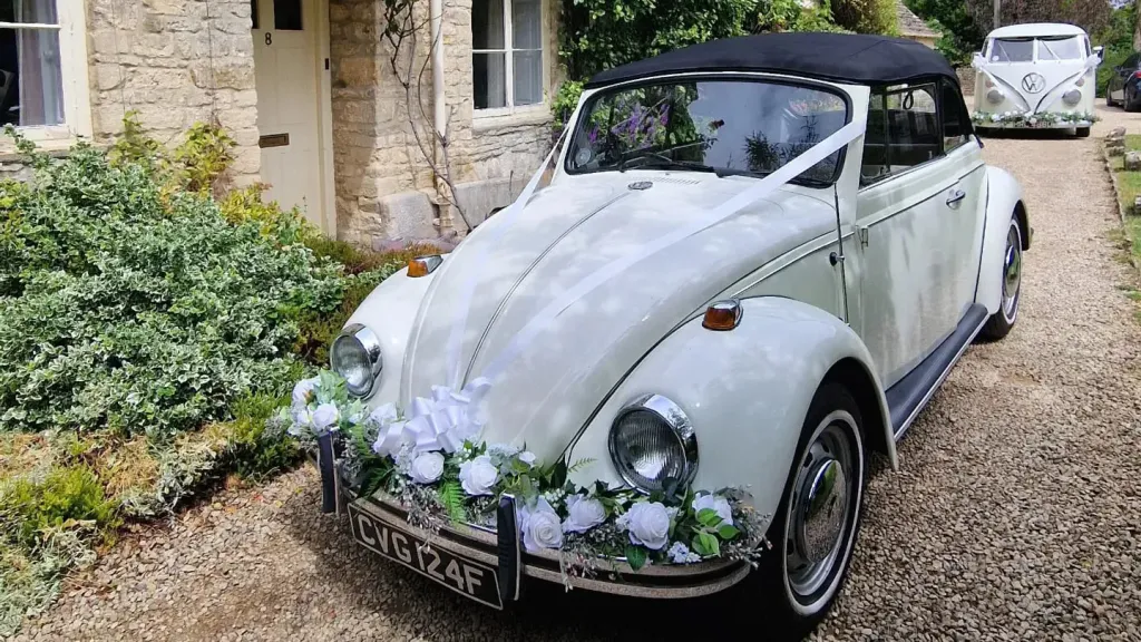 White Classic VW Beetle with soft top black roof parked in front of wedding venue in Cotswolds. Car is decorated with Green foliage on its front chrome bumper and white ribbons accross its bonnet. Campervan in far background