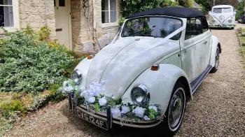 White Classic VW Beetle with soft top black roof parked in front of wedding venue in Cotswolds. Car is decorated with Green foliage on its front chrome bumper and white ribbons accross its bonnet. Campervan in far background