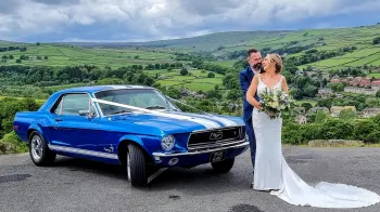 Classic Blue Ford Mustang dressed with White Ribbons across its bonnet. Bride and Groom standing in front of the car holding each others.