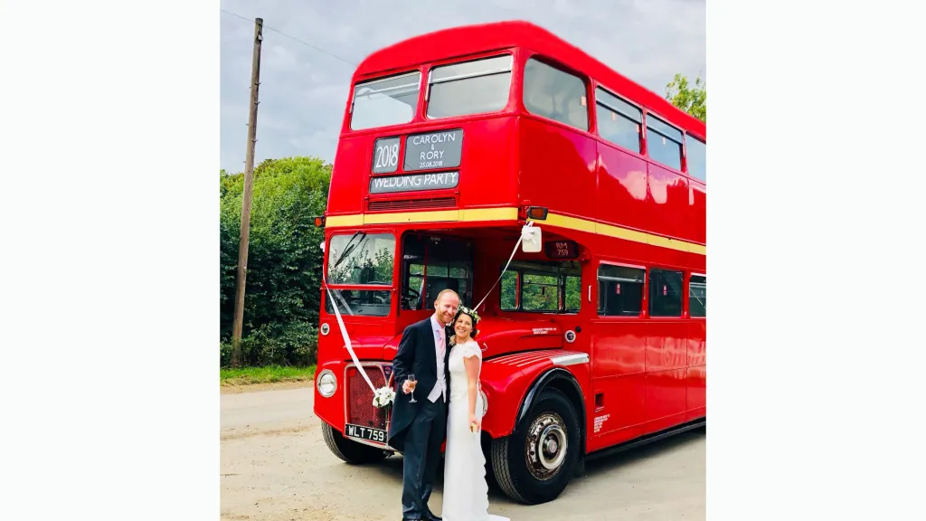 Bride and Groom standing on front of a vintage Red Routemaster Wedding bus decorated with white wedding ribbons
