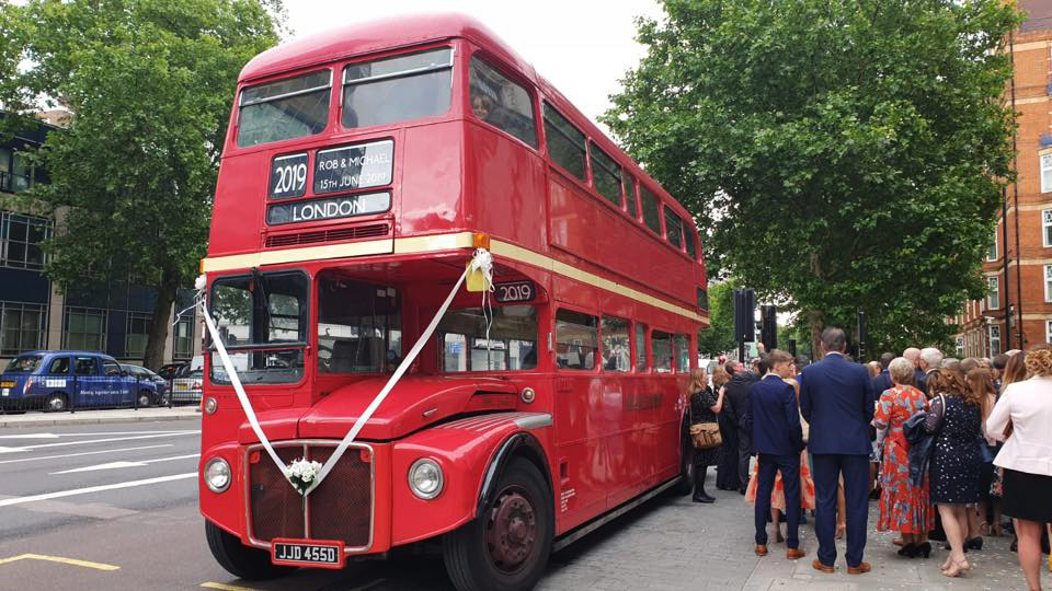 Big red double decker London Routemaster Bus decorated white white ribhbons with wedding guests queuing to board the bus
