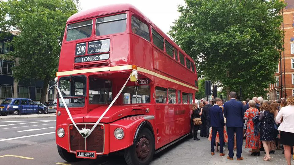 Big red double decker London Routemaster Bus decorated white white ribhbons with wedding guests queuing to board the bus