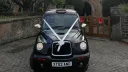 Front view of black taxi cab with white wedding ribbons and headlights on