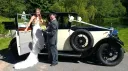 Bride wearing a white wedding dress exiting a black and cream car with the help of the chauffeur wearing a full uniform and hat.