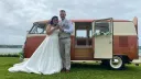 Bride and Groom standing in front of a classic VW Campervan in two tone Orange and white with rear passenger door open