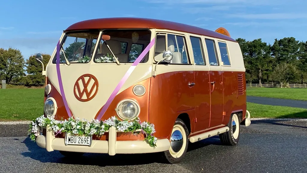 Classic VW Splitscreen campervan in two tone White and burnt orange decorated with green foliage on front bumper and white wedding ribbons