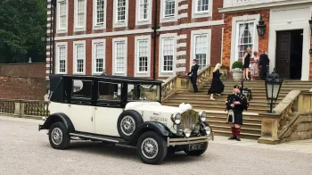 Ivory imperial wedding car with black wheel arches and roof parked in front of a wedding venue in Liverpool. Bagpipe player in the background with some wedding guests