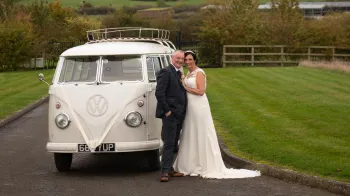 Bride wearing a white dress standing in front of a Classic Campervan decorated with white Ribbons with her Groom