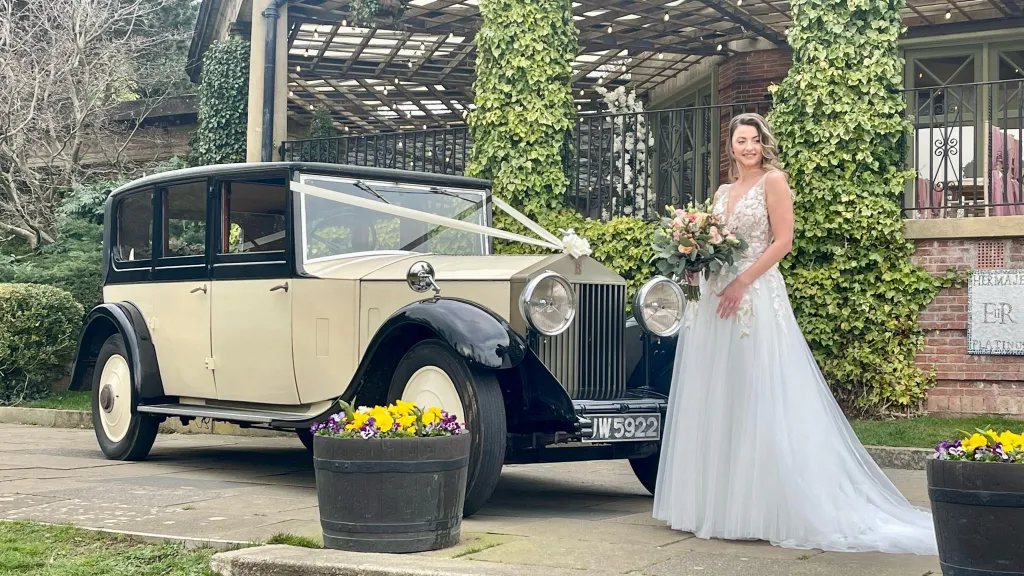 Bride in a white dress holding a bouquet of flowers standing in front of a Vintage Rolls-Royce Limousine in Black and Ivory decorated with Ivory Ribbons across front bonnet
