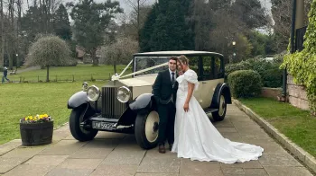 Bride and Groom standing in front of a vintage Rolls-Royce dressed with Ivory ribbons and bows. Bride is wearing a white wedding dress