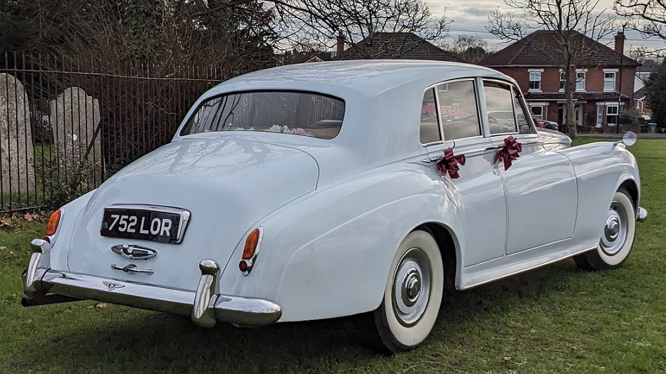 Rear view of a classic Bentley with large white wall tires and Burgundy bows on door handles