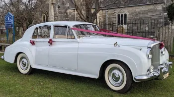 Right side view of a classic Bentley in White parked on the grass in front of a church in Portsmouth dressed with Burgundy ribbons and bows