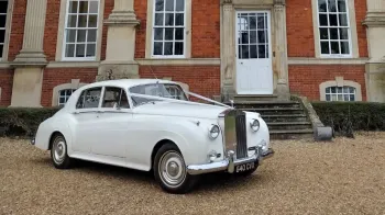 Front right side view of a white Rolls-Royce silver Cloud with white ribbons parked outside a wedding venue
