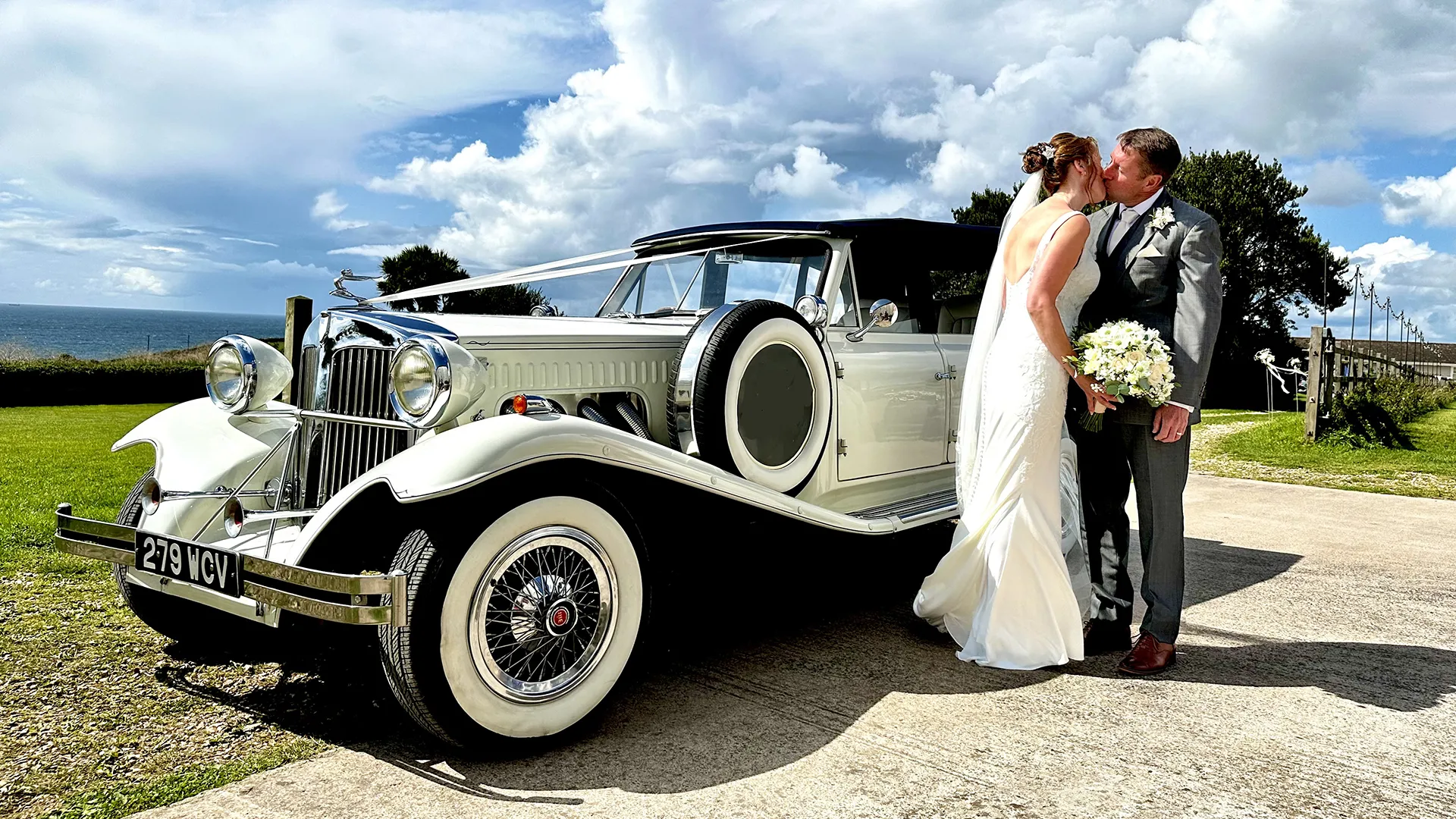 Trendy vintage-style white Beauford wedding car with large white wall tires with the coastline in the background and a Bride wearing a white wedding dress and Groom kissing next to it under summer sky.