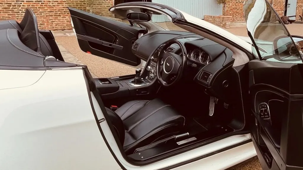 White Aston Martin with convertible roof open and both passenger and driver's doors open showing a black leather interior