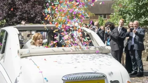 Bride and Groom seating inside a Classic Daimler Limousine with Roof open looking backward while the Confetti Cannons installed inside the car have been fired. Wedding Ghuests cheering in the background.