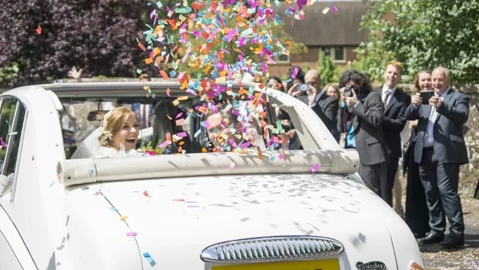 Bride and Groom seating inside a Classic Daimler Limousine with Roof open looking backward while the Confetti Cannons installed inside the car have been fired. Wedding Ghuests cheering in the background.