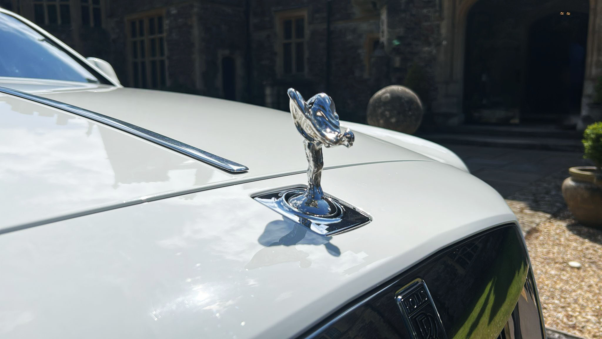 Close-up of the Spirit of Ecstasy bonnet ornament on a white Rolls-Royce Ghost, with sunlight reflecting off the polished chrome and historic stone architecture blurred in the background.