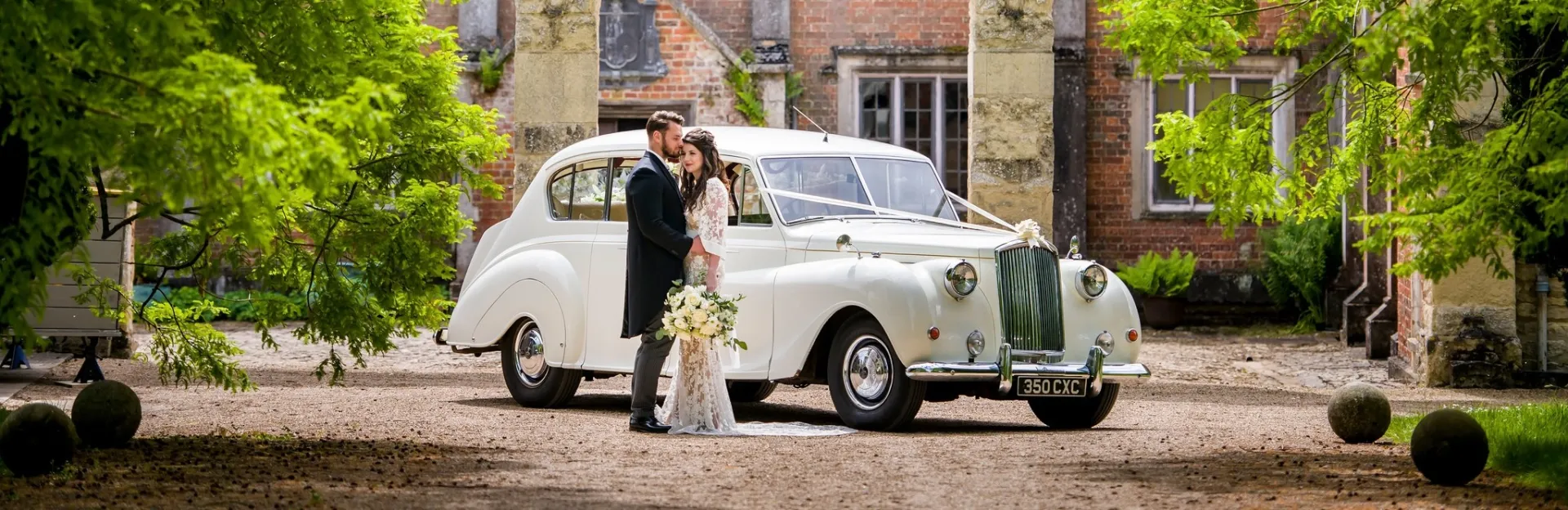 Bride and groom standing in front of a Classic white vintage wedding car decorated with ivory ribbons and bows under a stone archway, surrounded by greenery and a historic red-brick manor house in the background