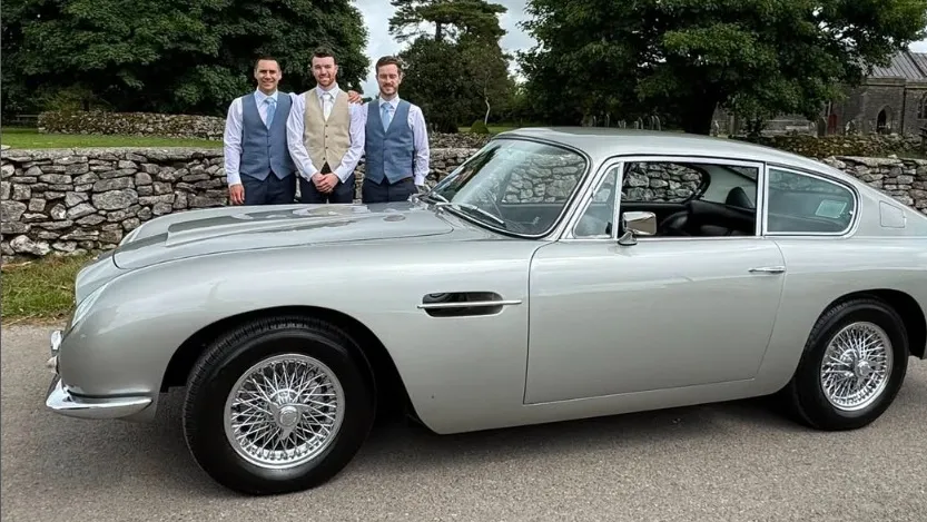 Left side view of a Silver Classic Aston Martin DB6 with Groom and his two groomsmen standing behing the vehicle