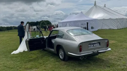 Classic aston Martin DB6 in Silver with front passenger door open. Bride and Groom walking towards the Marquee in the background