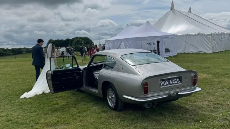 Classic aston Martin DB6 in Silver with front passenger door open. Bride and Groom walking towards the Marquee in the background