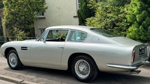 Left side view of a classic Aston Martin DB6 with chrome spokes wheels parked in the side of the road with greenery in the background