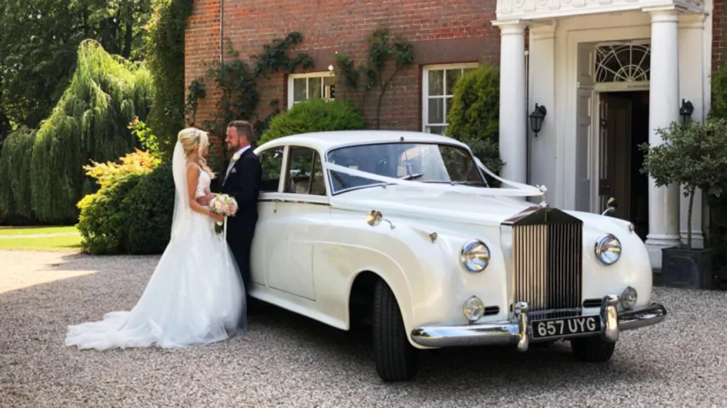 Bride and Groom holding each others standing by a classic white rolls-royce silver cloud with a manor house style wedding venue in the background