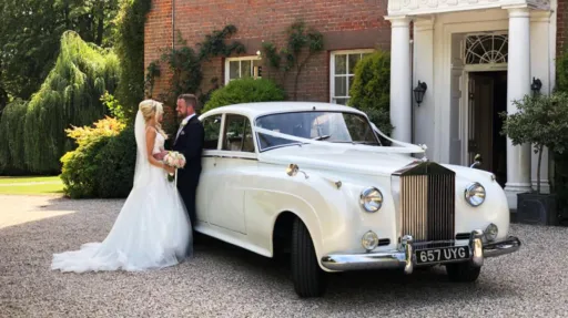 Bride and Groom holding each others standing by a classic white rolls-royce silver cloud with a manor house style wedding venue in the background