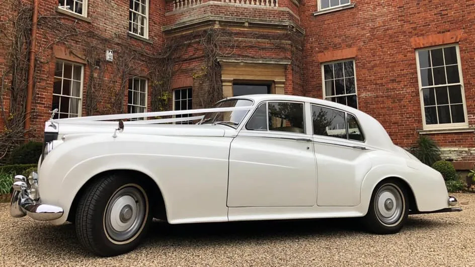 Left side view of white Classic Rolls-Royce Silver Cloud with white wedding ribbons, thin white wall tires, parked in front of a red brick manor house style wedding venue