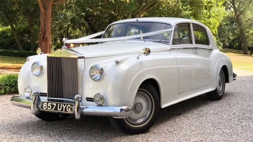 White Classic Rolls-Royce Silver Cloud decorated with Ivory wedding ribbons, thin white wall tires, large chrome grill and front bumper with greenery in the backgroun