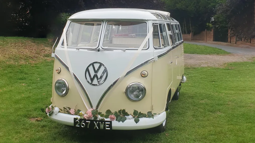 Front view Classic Volkswagen Campervan in White & Cream with garland of pink flowers on front bumper and light pink ribbons accros the front of its bonnet