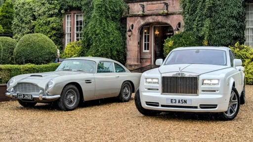 A classic silver Aston Martin DB5 parked next tp a modern white Rolls-Royce Phantom, both displayed on a gravel driveway in front of an elegant ivy-clad manor house.