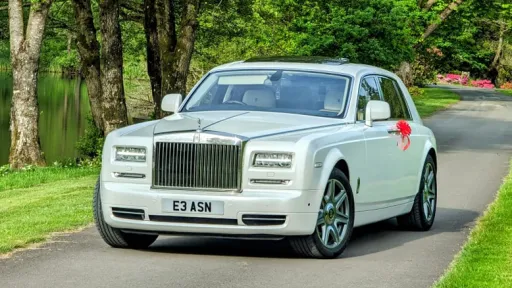 Modern White Rolls-Royce Phantom with red wedding bows on door handle parked in the middle of a tarmac path with green forest in the background