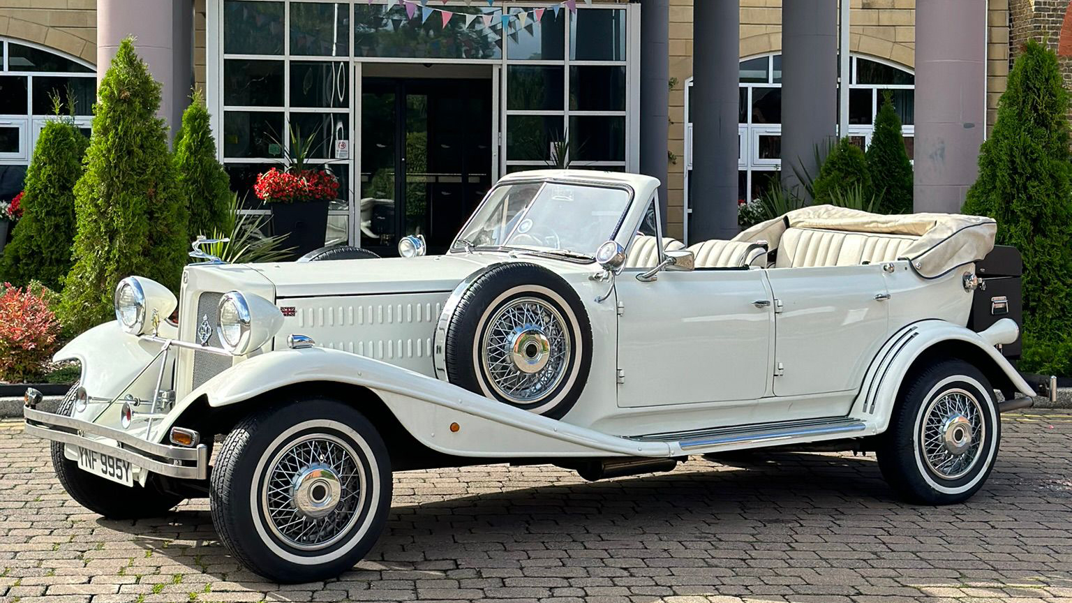 White Vintage-style Beauford with Convertible roof down, thin white wall tires and spare wheel mounted in the side of the vehicle