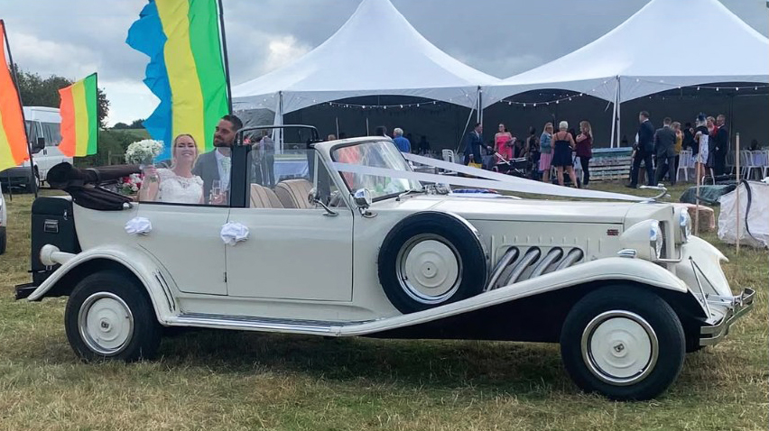 Beauford convertible parked in front of a wedding marquee with Bride and Groom seating inside the vehicle smiling.