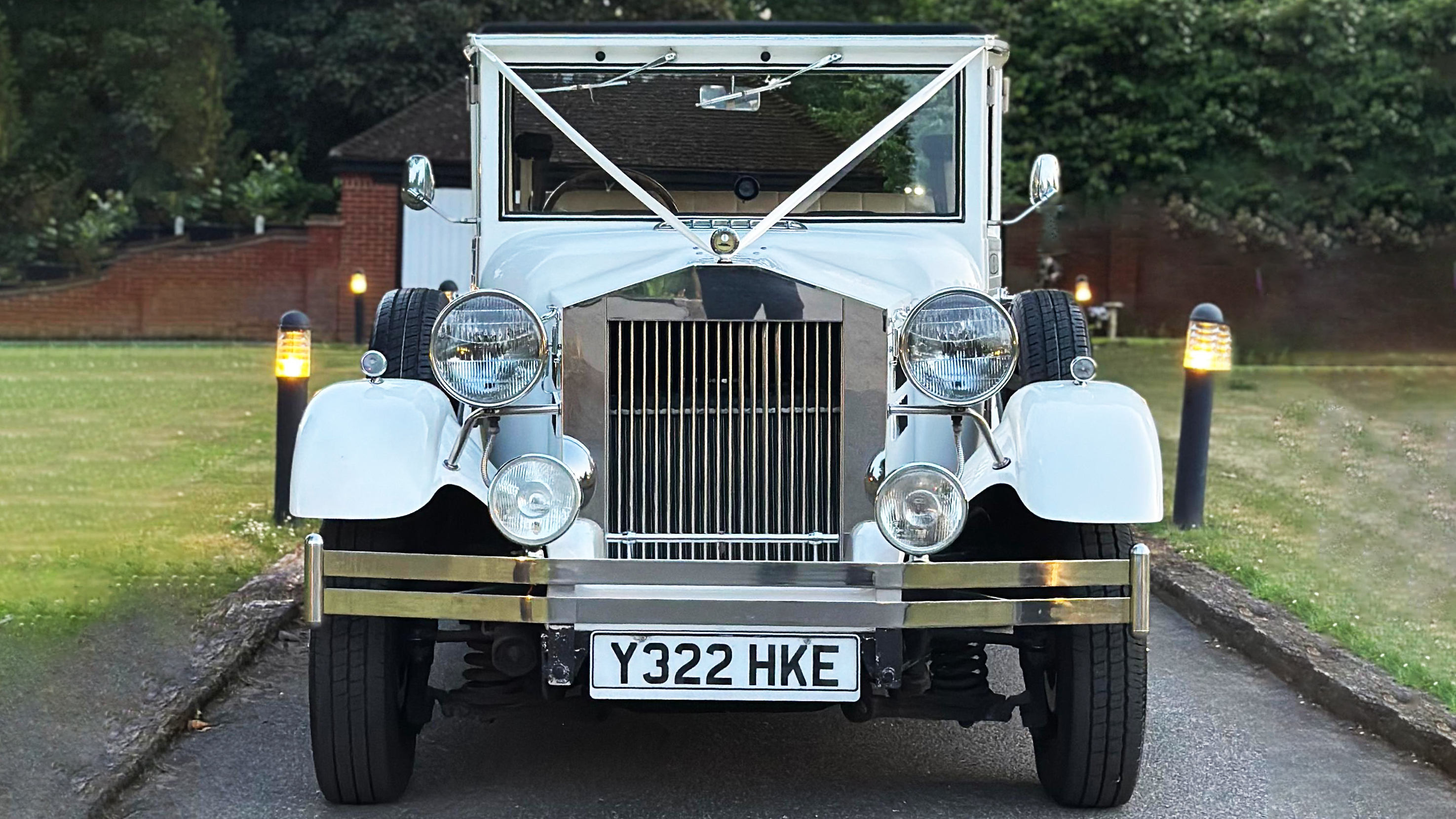 Full front view of white imperial wedding car with white ribbons across front bonnet, large front chrome grill. The vehicle is parked in the middle of a path with greenery on both side