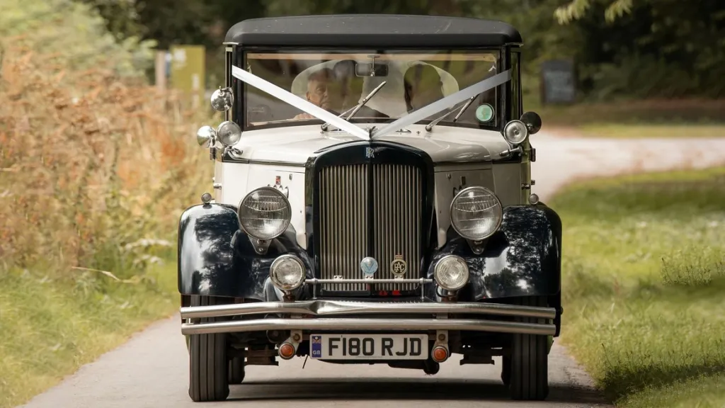 Full front view of vintage style regent dressed with white ribbons across its front bonnet behing driven on a small road. passengers and drivers can be seen inside the vehicle