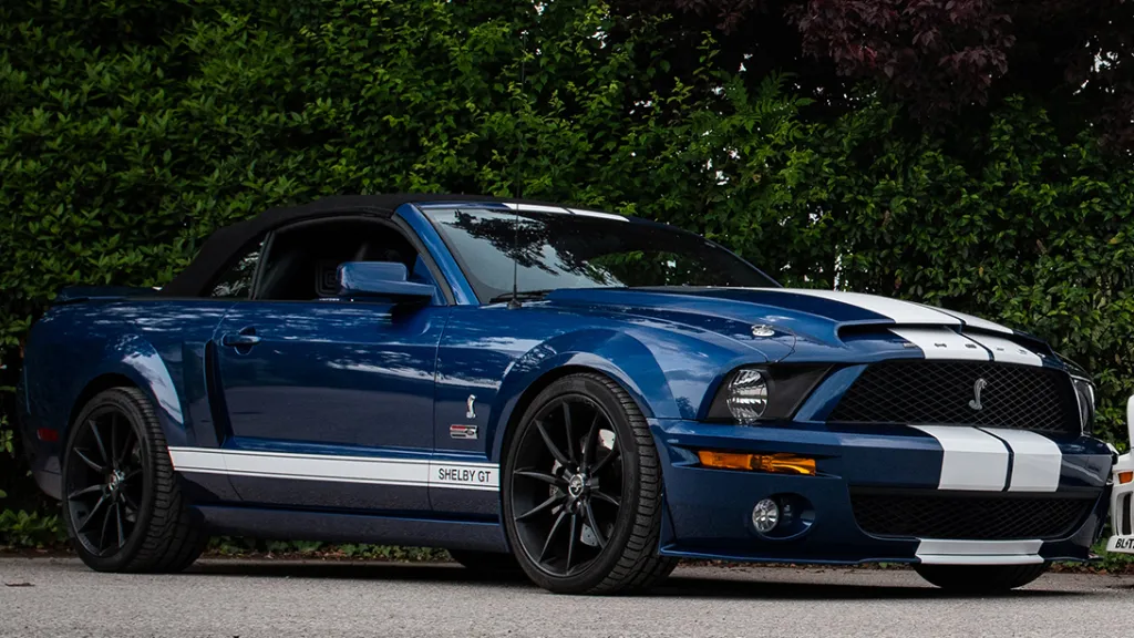 Blue ford Mustand Shelby with white stripes, black alloy wheels and black soft-top convertible rood