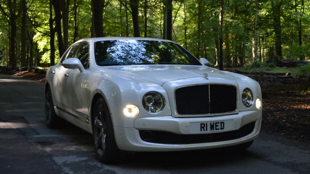 White Bentley Mulsanne parked in the middle of a path in the forest with front headlights on