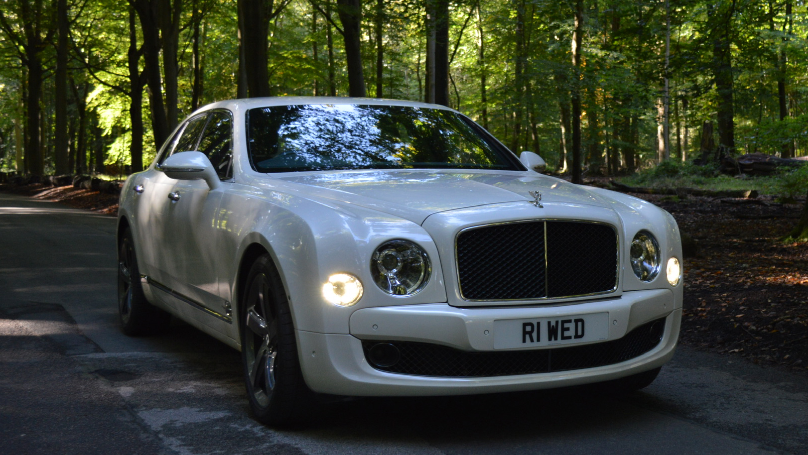 White Bentley Mulsanne parked in the middle of a path in the forest with front headlights on