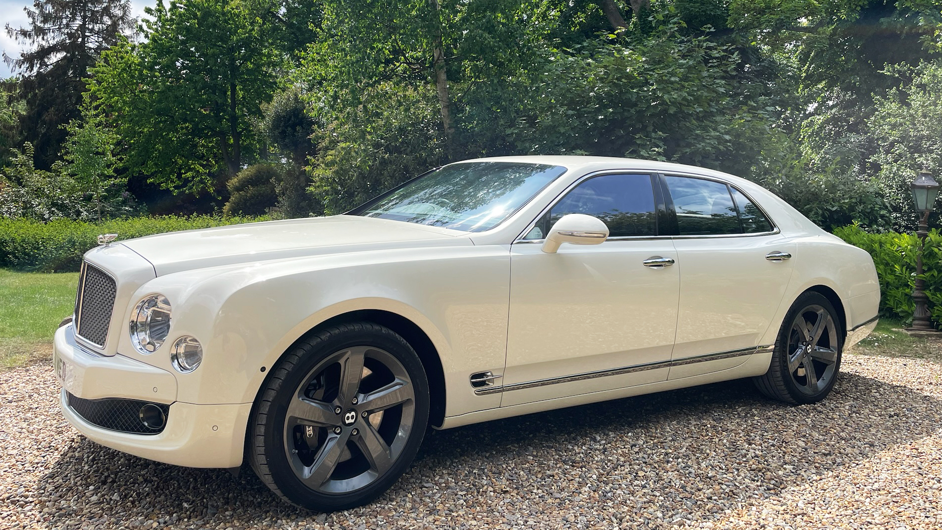 Left side view of White Bentley Mulsanne with silver alloy wheels. Car is parked on a gravel path with greenery in the background