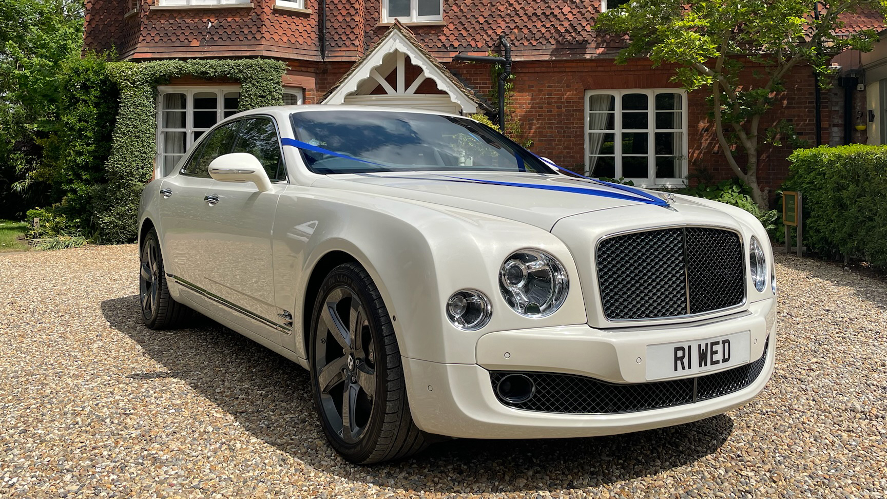 White Bentley Mulsanne decorated with royale blue ribbons across the front bonnet and red brick wall wedding venue in the background
