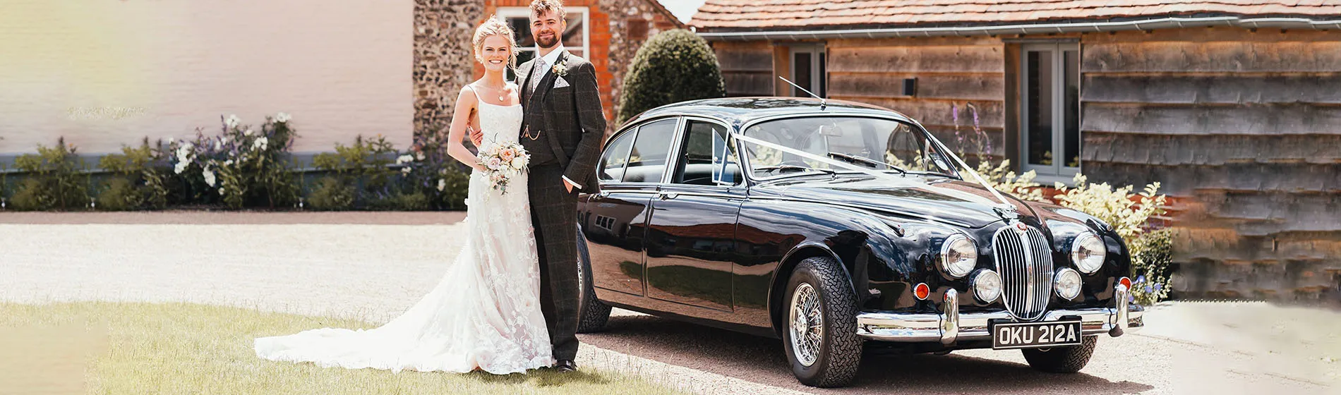 Bride and groom posing next a classic black Jaguar wedding car decorated with white ribbon, parked outside a rustic barn venue on their wedding day.