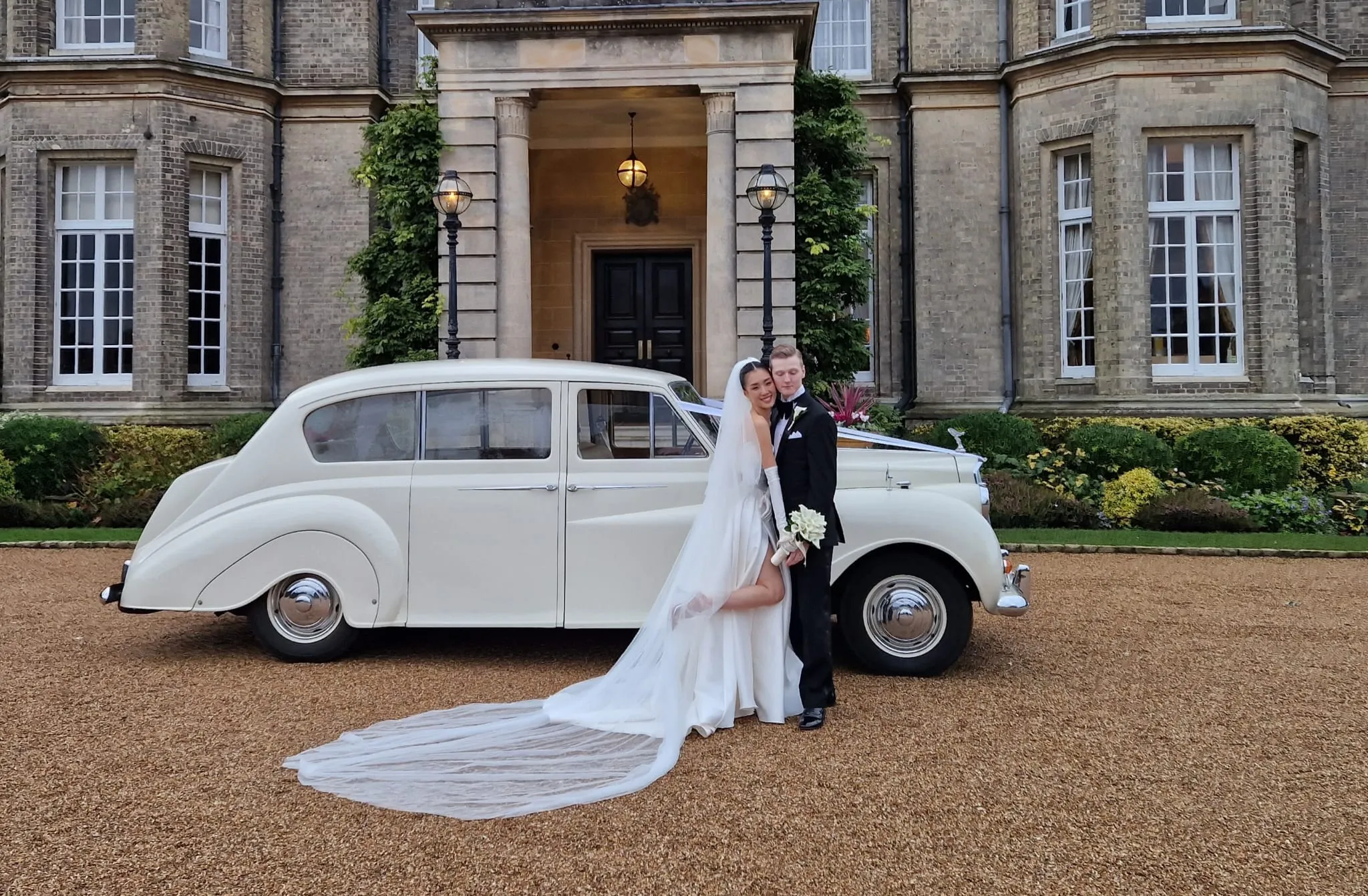 White classic Wedding Car decorated with white ribbons parked in front of a wedding venue with Bride and Groom standing and holdign each others in front of the vehicle