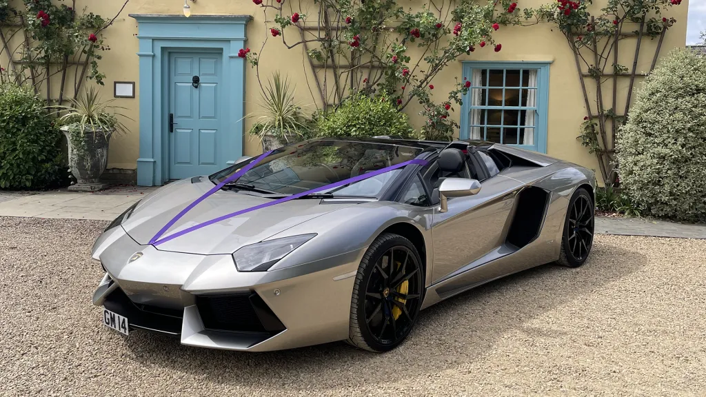 Silver Lamborghini Aventador with purple wedding ribbons, parked in front of a quaint country house