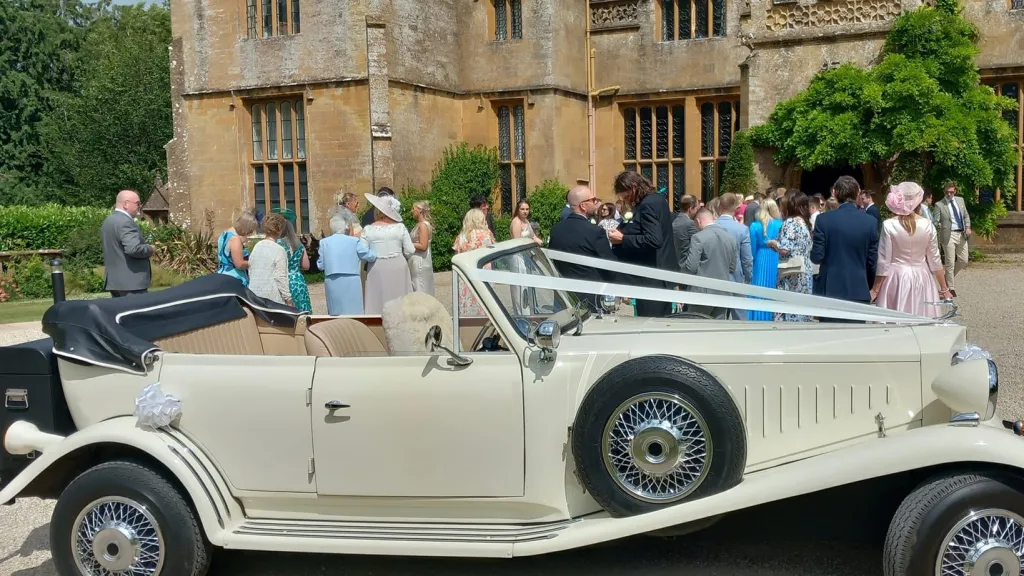Right Side view of Ivory Beauford with Roof down, Spare wheel mounted on right wheel arch and Traditional V-shape white ribbon accross the bonnet. Wedding guests are stangin in the background
