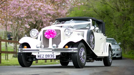 Full front view of a Vintage Beauford dressed with pink ribbons and a large bow in being driven on the road. pink blossom tree in the background