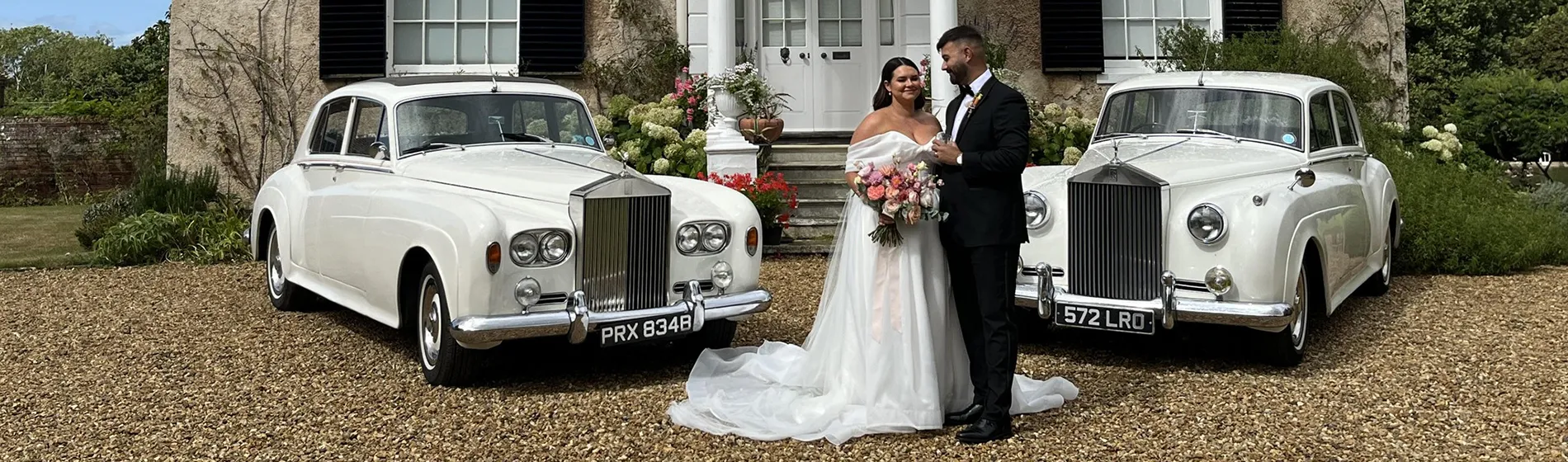 Two Classic Rolls-Royce Silver Clouds in White with Bride and Groom standing in the middle of both vehicles parked on gravel in front of a stately home.